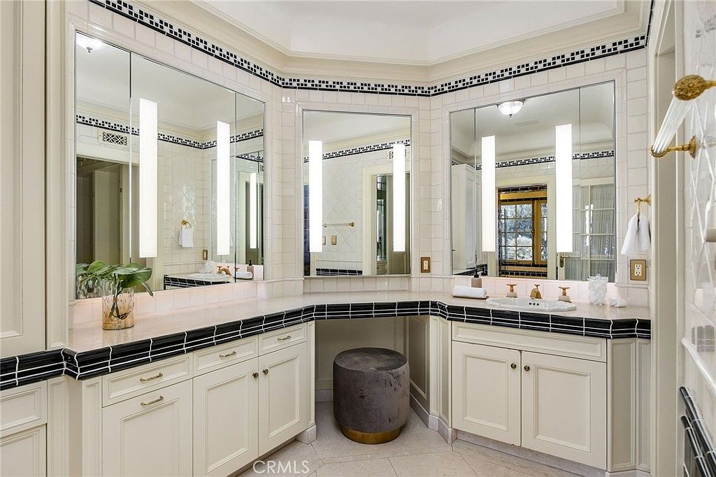 This elegant primary bathroom features a dual-vanity setup with light-toned cabinetry and a central makeup vanity area. The space is accented by a decorative black and white tile border that runs along the wall and countertop edges, complemented by large mirrors and integrated vertical lighting. A soft, neutral-toned stool sits beneath the vanity, creating a sophisticated and functional retreat.