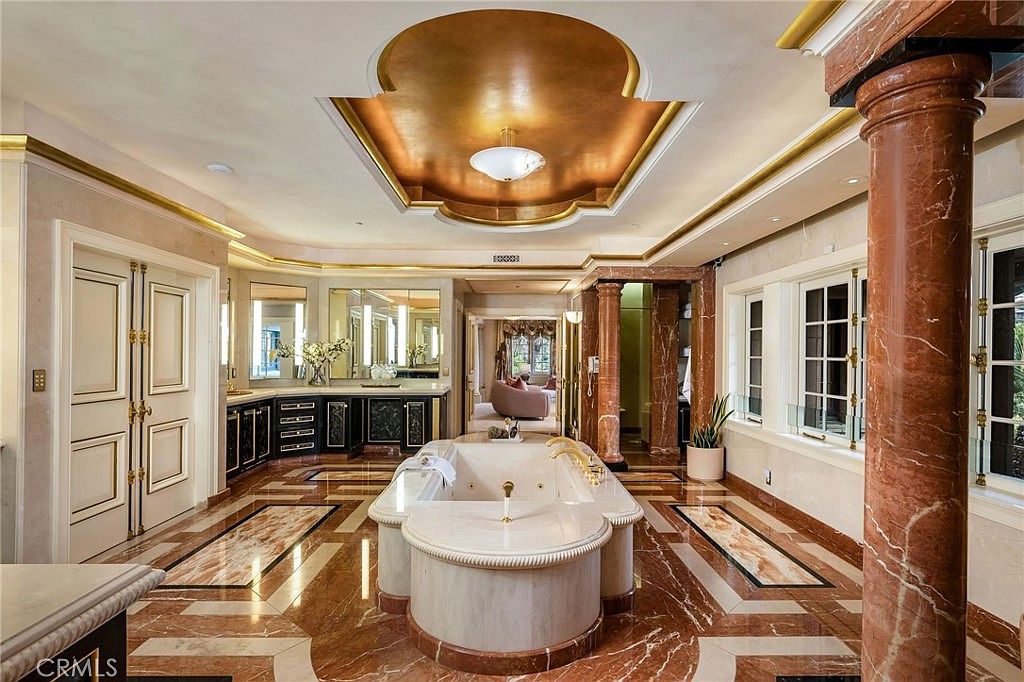 This opulent primary bathroom features a central soaking tub encased in stone, set against a backdrop of rich, reddish-brown patterned stone flooring and matching columns. The room is crowned by a dramatic, gold-toned recessed ceiling feature with a central light fixture, while a vanity area with dark cabinetry and mirrors lines the back wall. The perspective is a wide, symmetrical shot from the entrance, emphasizing the grand scale and luxurious, classic design of the space.