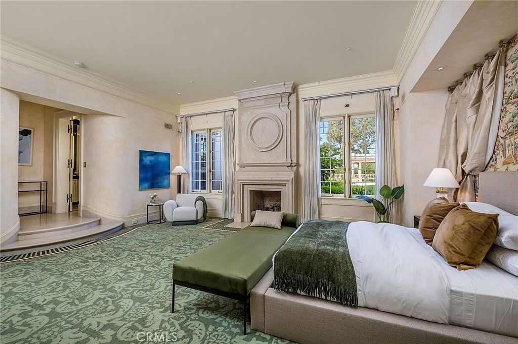 This spacious primary bedroom features a large bed with a neutral-toned headboard, complemented by a long green bench at the foot. A prominent stone-clad chimney breast serves as a focal point between two sets of tall windows that allow for ample natural light. The room is finished with patterned wall-to-wall carpeting and a sophisticated, traditional aesthetic.
