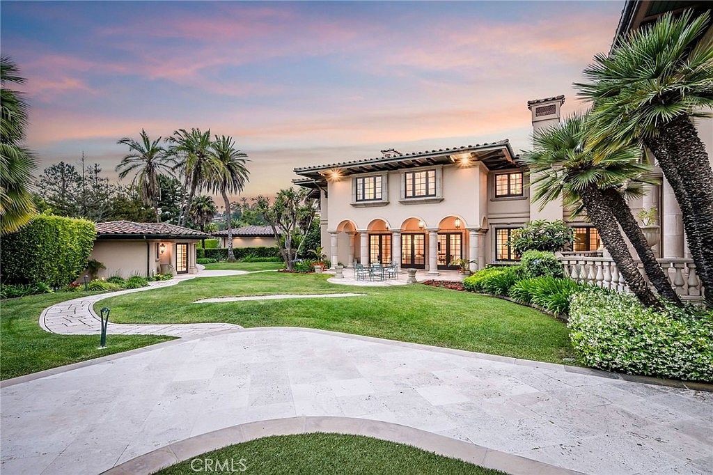 This expansive rear view of a Mediterranean-style estate showcases a large, manicured lawn leading to a multi-story residence with arched loggias and a tiled roof. The property features a stone-paved patio area, mature palm trees, and a separate smaller structure to the left, all set against a soft, sunset-lit sky. The perspective is captured from the ground level, emphasizing the grand scale and serene outdoor living space of the home.
