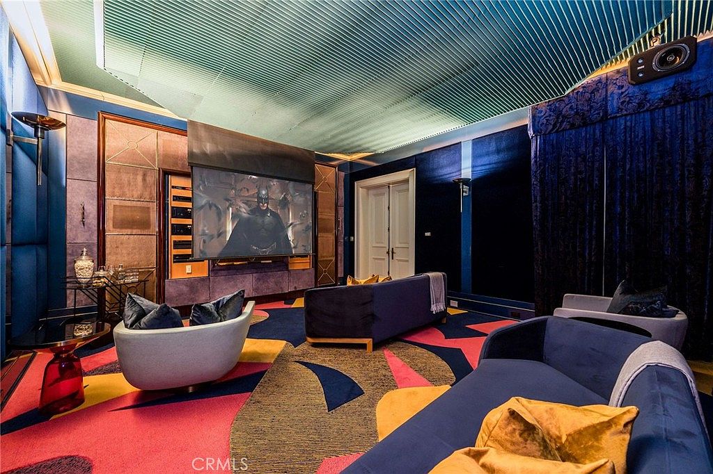 This image showcases a vibrant and theatrical home theater room featuring a large projection screen displaying a movie. The space is furnished with plush blue sofas and a unique, colorful patterned carpet, while the ceiling is finished with a textured, ribbed design. The room is enclosed by dark blue walls and heavy curtains, creating an immersive, cinematic atmosphere.