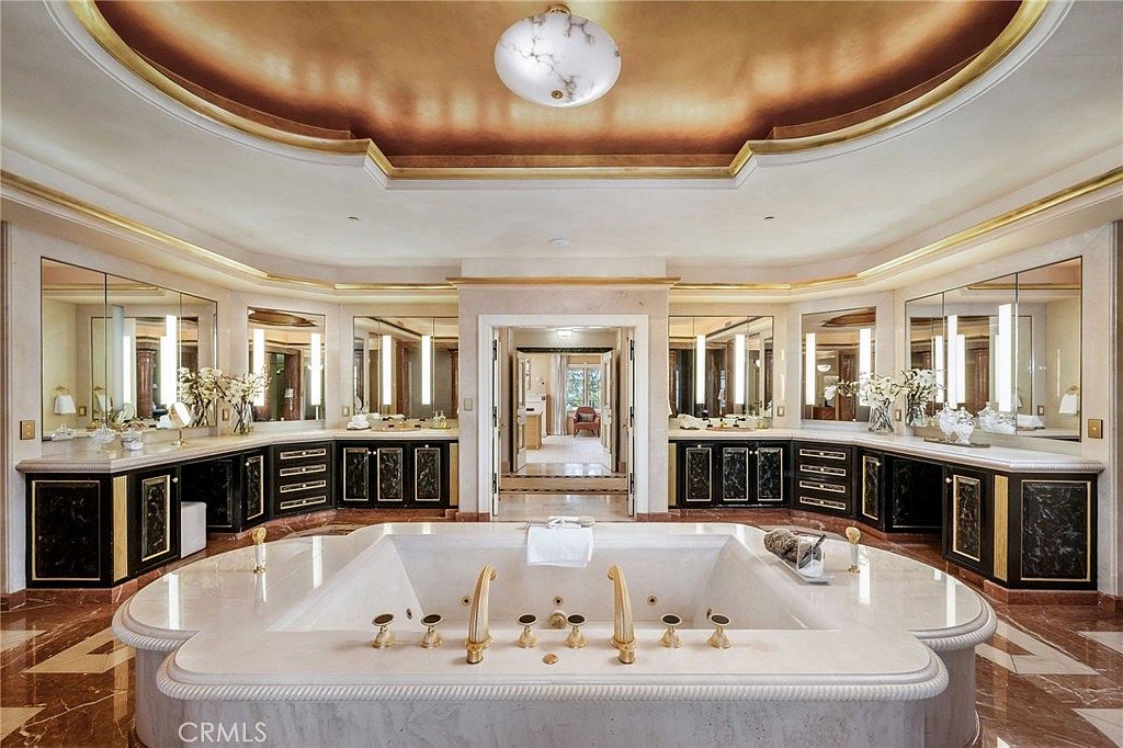 This opulent primary bathroom features a large, central soaking tub with gold-toned fixtures as the focal point. The room is symmetrically designed with dual vanity areas, extensive mirror work, and a warm, metallic-toned ceiling treatment. The space is finished with polished stone flooring and dark cabinetry, creating a luxurious and formal atmosphere.