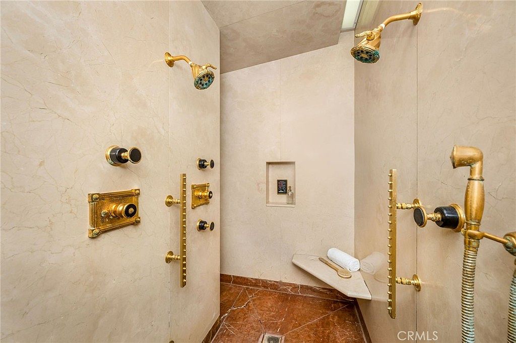 This spacious walk-in shower features light-toned stone wall tiles and a contrasting reddish-brown patterned tile floor. The shower is equipped with multiple gold-toned fixtures, including two overhead showerheads, a handheld sprayer, and various wall-mounted controls, alongside a built-in stone bench and a recessed wall niche for storage. The perspective is from the entrance of the shower, showcasing the extensive plumbing and luxurious, spa-like design.