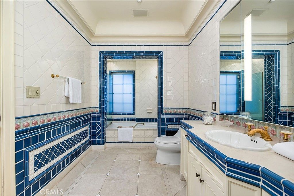 This primary bathroom features a distinctive Mediterranean-inspired design with bold blue and white tile work throughout. The space includes a vanity with a scalloped sink, a separate toilet area, and a bathtub set within a tiled alcove. The room is brightened by a large mirror and recessed lighting, creating a cohesive and classic aesthetic.