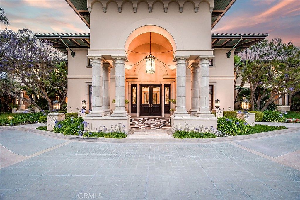 This grand entryway features a prominent arched portico supported by four substantial stone columns, creating a stately and welcoming focal point. The double front doors are framed by dark wood and glass, leading to a patterned stone walkway that extends into a circular driveway. Lush landscaping with vibrant green shrubs and purple flowering plants softens the architectural lines, while decorative lanterns provide elegant lighting against the light-toned stucco facade.