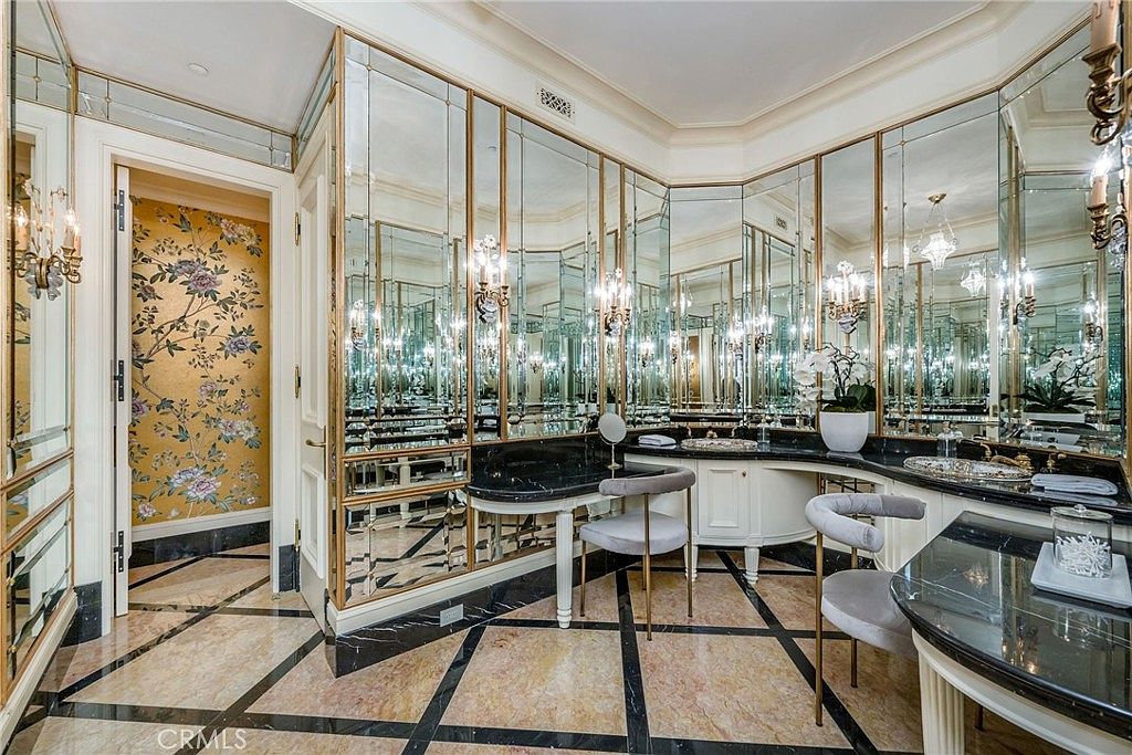 This opulent primary bathroom features an expansive vanity area with dual sinks set into dark stone countertops and a dedicated makeup station. The walls are adorned with floor-to-ceiling mirrored panels framed in gold, creating a dramatic and reflective atmosphere, while the floor showcases intricate patterned stone tile. A doorway leads to an adjacent space decorated with vibrant floral wallpaper, adding a touch of classic elegance to the luxurious design.