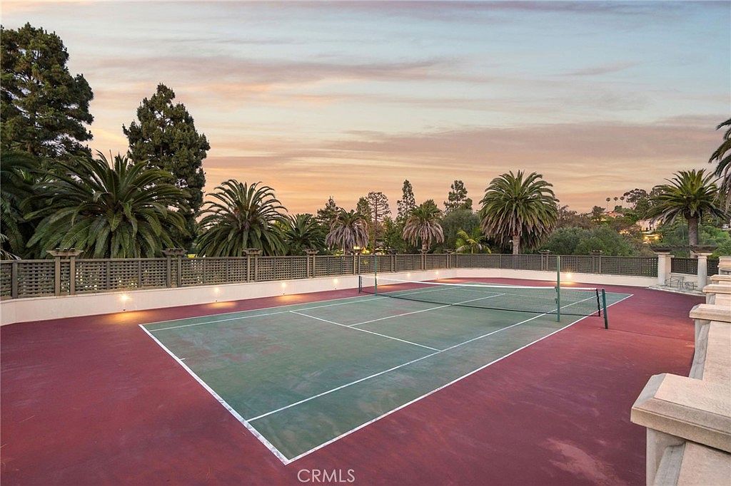This image features a private tennis court set against a scenic sunset sky with soft, warm lighting. The court is enclosed by a decorative lattice-style fence and is surrounded by lush palm trees and mature landscaping, creating a serene and exclusive atmosphere. The perspective is captured from an elevated vantage point, highlighting the expansive layout and the tranquil outdoor setting.