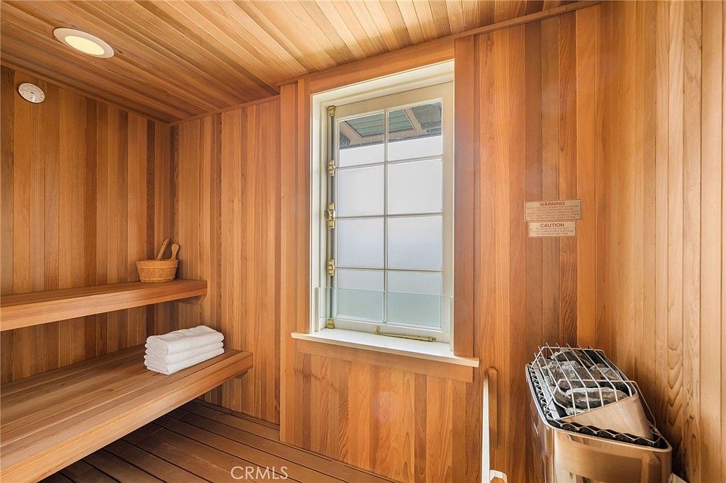 This image showcases a private home sauna constructed with warm-toned wood paneling on the walls and ceiling. The space features a built-in wooden bench, a stack of clean white towels, a wooden bucket, and a metal heater filled with stones. A window with frosted glass provides natural light, creating a serene and relaxing atmosphere.