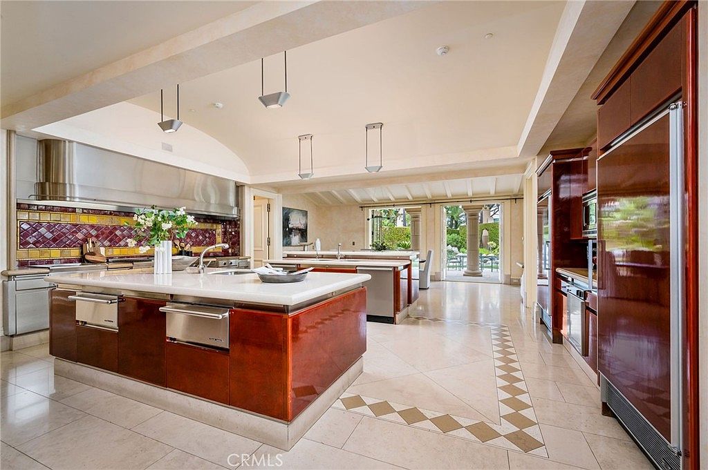 This expansive, high-end kitchen features a large central island with a light-toned stone countertop and a glossy, dark-toned base. The space is defined by a dramatic, curved ceiling design with recessed lighting and a prominent stainless steel range hood above a vibrant, multi-colored tiled backsplash. The open-concept layout flows into a secondary prep area and leads toward a bright, sunlit transition space with large glass doors opening to the outdoors.