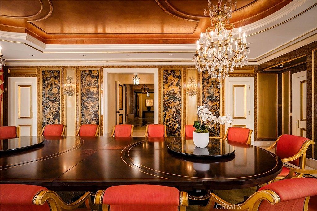 This opulent dining room features a large, dark-toned oval table surrounded by numerous chairs upholstered in vibrant red fabric. The space is defined by ornate gold-trimmed wall panels with intricate dark patterns, a coffered ceiling with a warm metallic finish, and a grand crystal chandelier as the central focal point. The perspective is centered, looking through the dining area toward a bright, marble-floored hallway that leads to another room, creating a sense of formal elegance and luxury.