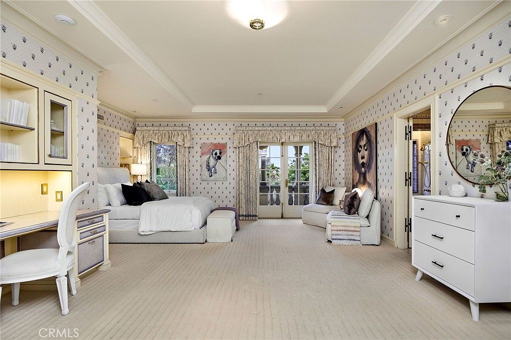 This spacious primary bedroom features a neutral color palette with patterned wallpaper and light-toned carpeting. The room includes a built-in desk area, a plush bed with dark accent pillows, and a cozy seating area near the glass doors that lead to a balcony. A large white dresser with a circular mirror adds a bright, functional touch to the space, creating an airy and elegant atmosphere.