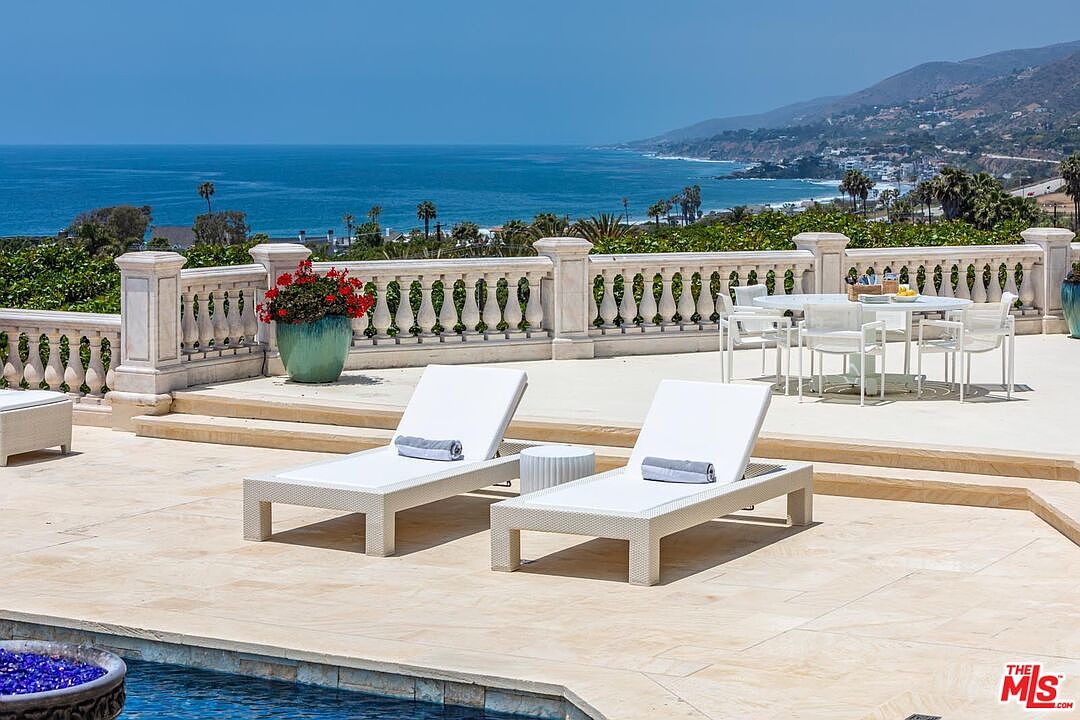 This luxurious outdoor terrace features a spacious stone deck equipped with modern white lounge chairs and a dining set, all overlooking a breathtaking panoramic view of the coastline and ocean. The area is framed by an elegant stone balustrade and a vibrant potted plant, creating a sophisticated and serene atmosphere perfect for relaxation or entertaining. The perspective captures the seamless transition between the high-end patio furniture and the expansive natural beauty of the horizon.