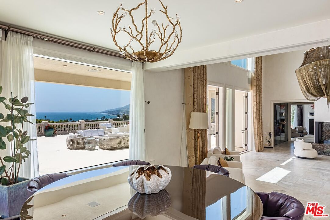 This elegant dining room features a polished circular table topped with a decorative shell-shaped centerpiece, set beneath a striking, organic-shaped gold chandelier. The space seamlessly transitions to a sprawling outdoor terrace through floor-to-ceiling glass doors, offering a breathtaking, unobstructed view of the ocean and coastline. The interior design is sophisticated and airy, with neutral tones and high-end finishes that emphasize the luxurious indoor-outdoor living experience.