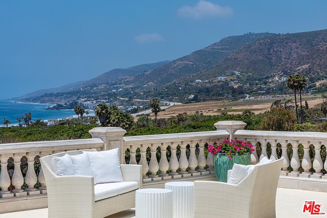 This elevated balcony offers a breathtaking, panoramic view of the Pacific coastline, featuring a classic stone balustrade that frames the scenic landscape. The space is furnished with comfortable white outdoor seating and a vibrant potted plant, creating an inviting atmosphere for relaxation. The perspective captures the seamless blend of luxury outdoor living with the natural beauty of the ocean and rolling hills in the distance.