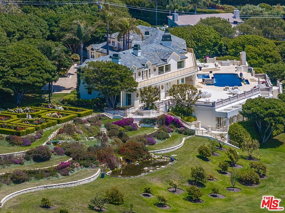 This high-angle aerial view captures a sprawling, luxurious estate featuring a grand multi-story mansion with a slate roof and expansive terraces. The property boasts meticulously manicured formal gardens, a large swimming pool, and a naturalistic pond set within a lush, rolling landscape. The overall impression is one of opulence, privacy, and sophisticated architectural design.