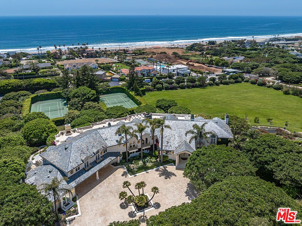 This stunning aerial view captures a sprawling, luxurious estate nestled along the coastline, featuring a large slate-roofed mansion with a grand circular driveway. The property boasts extensive grounds, including two private tennis courts and a vast, manicured lawn that stretches toward the ocean. The perspective highlights the home's impressive scale, architectural elegance, and its prime location with direct proximity to the beach.