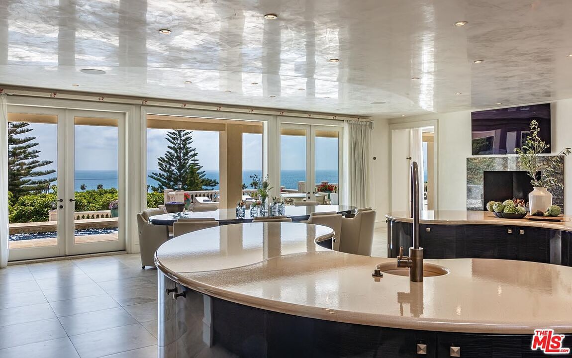 This luxurious, open-concept kitchen features a large, curved island with a smooth, light-colored countertop and dark cabinetry, serving as the focal point of the room. The space is highlighted by a high-gloss, reflective ceiling and expansive glass doors that offer a stunning, panoramic view of the ocean and outdoor terrace. The design seamlessly blends sophisticated indoor living with breathtaking coastal scenery, creating an airy and elegant atmosphere.