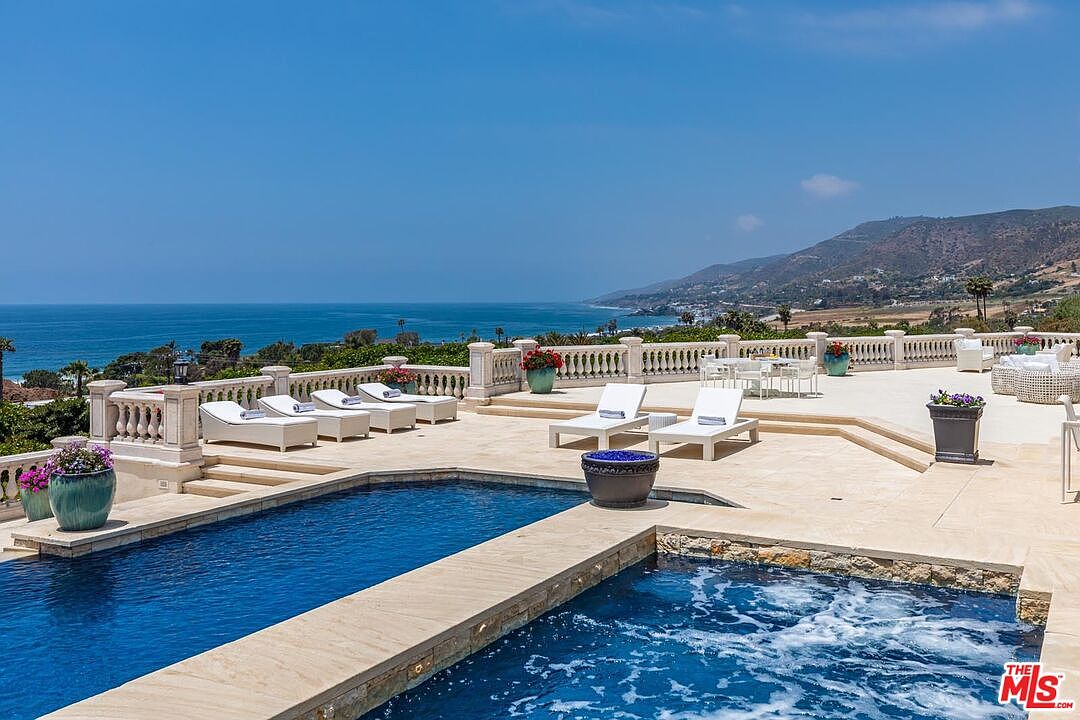 This stunning outdoor space features a luxurious infinity-edge pool and spa overlooking a panoramic view of the Pacific coastline. The expansive stone terrace is furnished with multiple white lounge chairs and elegant planters, creating a serene and sophisticated atmosphere perfect for relaxation. The cinematic perspective captures the seamless transition from the pristine pool deck to the vast blue horizon, highlighting the property's premier coastal location.