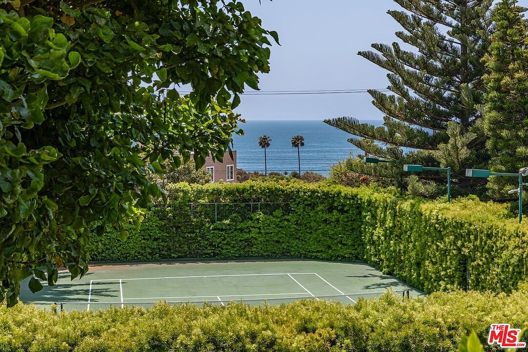 This elevated perspective captures a private tennis court nestled within lush, manicured hedges, creating a secluded recreational space. Beyond the court, the scene opens up to a breathtaking, unobstructed view of the sparkling blue ocean under a clear sky. The combination of active leisure space and serene coastal scenery provides a luxurious and tranquil atmosphere.