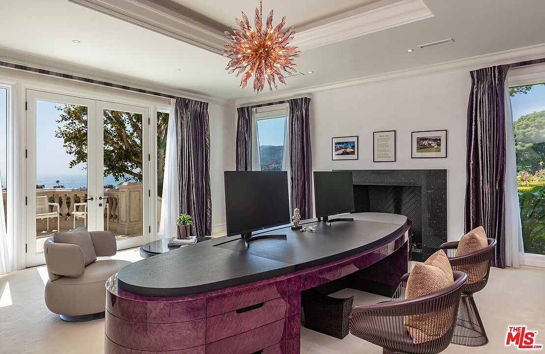 This luxurious home office features a striking, custom-designed purple high-gloss desk as its centerpiece, complemented by a dramatic starburst chandelier. The room is framed by elegant floor-to-ceiling purple curtains and offers a stunning, unobstructed view of the ocean through French doors leading to a balcony. A sleek fireplace and sophisticated decor create an atmosphere of refined productivity and high-end coastal living.