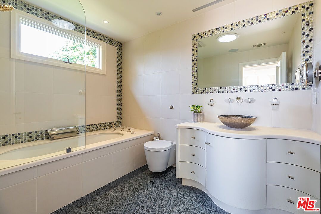 This modern primary bathroom features a sleek, curved white vanity with a unique vessel sink and a spacious soaking tub integrated with a glass shower partition. The room is accented by a decorative mosaic tile border that frames both the window and the large vanity mirror, creating a cohesive and stylish aesthetic. The dark, textured flooring provides a striking contrast to the bright white walls, resulting in a clean and contemporary atmosphere.