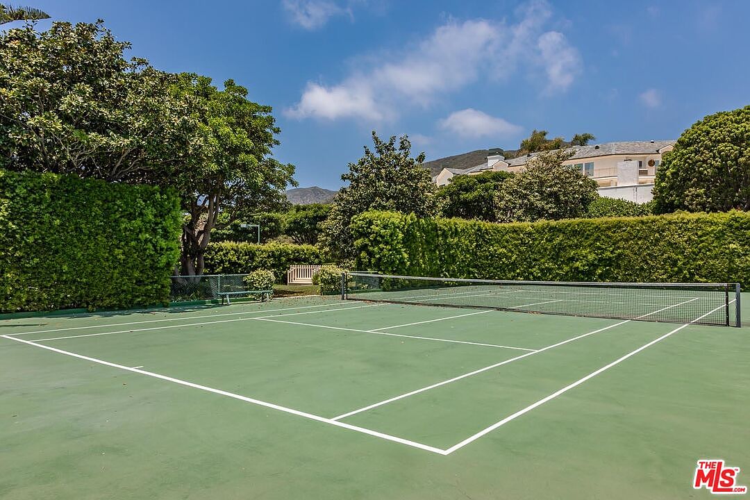 This image showcases a well-maintained private tennis court surrounded by lush, manicured hedges and mature trees. The perspective is from the court level, looking across the green surface toward the net and the distant hillside landscape. It presents an inviting, recreational space that suggests a high-end, active lifestyle.
