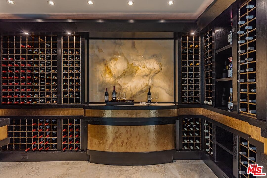 This sophisticated wine cellar features extensive custom dark wood cabinetry with integrated grid shelving for wine storage, flanking a central bar area. The focal point is a backlit, textured stone slab backsplash that provides a warm, luxurious glow, complemented by a hammered gold-toned metal bar counter. The perspective is a centered, eye-level shot that emphasizes the symmetry and high-end craftsmanship of the space.