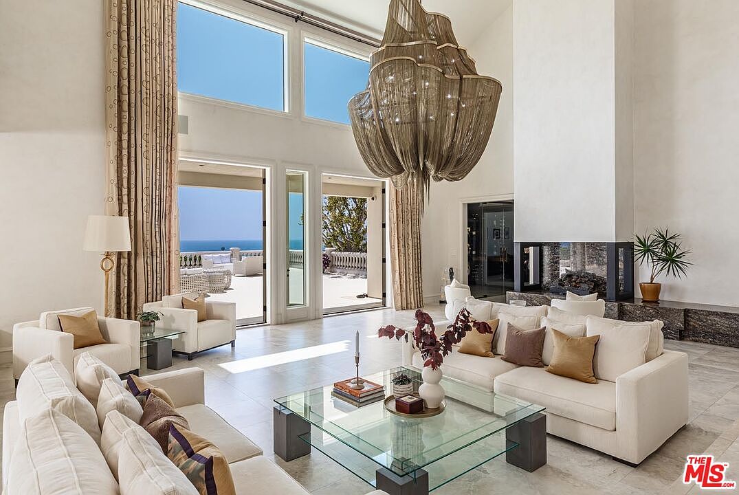 This expansive, high-ceilinged living room exudes luxury with its neutral color palette, plush cream sofas, and a striking, oversized metallic chandelier as the focal point. Large floor-to-ceiling windows and glass doors frame a breathtaking ocean view, seamlessly blending indoor comfort with the coastal landscape. The space is anchored by a modern fireplace and a glass-topped coffee table, creating an airy, sophisticated atmosphere perfect for high-end entertaining.