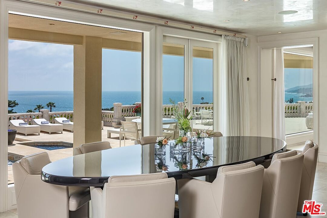 This elegant dining room features a sleek, oval-shaped black lacquer table surrounded by modern, cream-colored upholstered chairs. The room is framed by expansive floor-to-ceiling glass doors that open onto a sun-drenched terrace, offering a breathtaking, unobstructed view of the ocean. The bright, airy atmosphere is enhanced by a polished ceiling and minimalist decor, creating a sophisticated space perfect for luxury coastal living.
