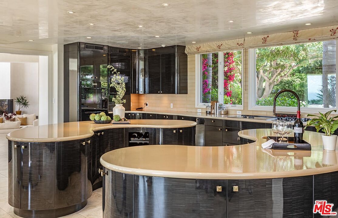 This sophisticated, high-end kitchen features a unique, sweeping curved island with a smooth, light-toned countertop that contrasts beautifully against the dark, high-gloss cabinetry. The room is illuminated by recessed lighting and large windows that provide a bright, airy feel while showcasing a lush green view outside. The overall aesthetic is modern and luxurious, emphasizing fluid lines and premium finishes.