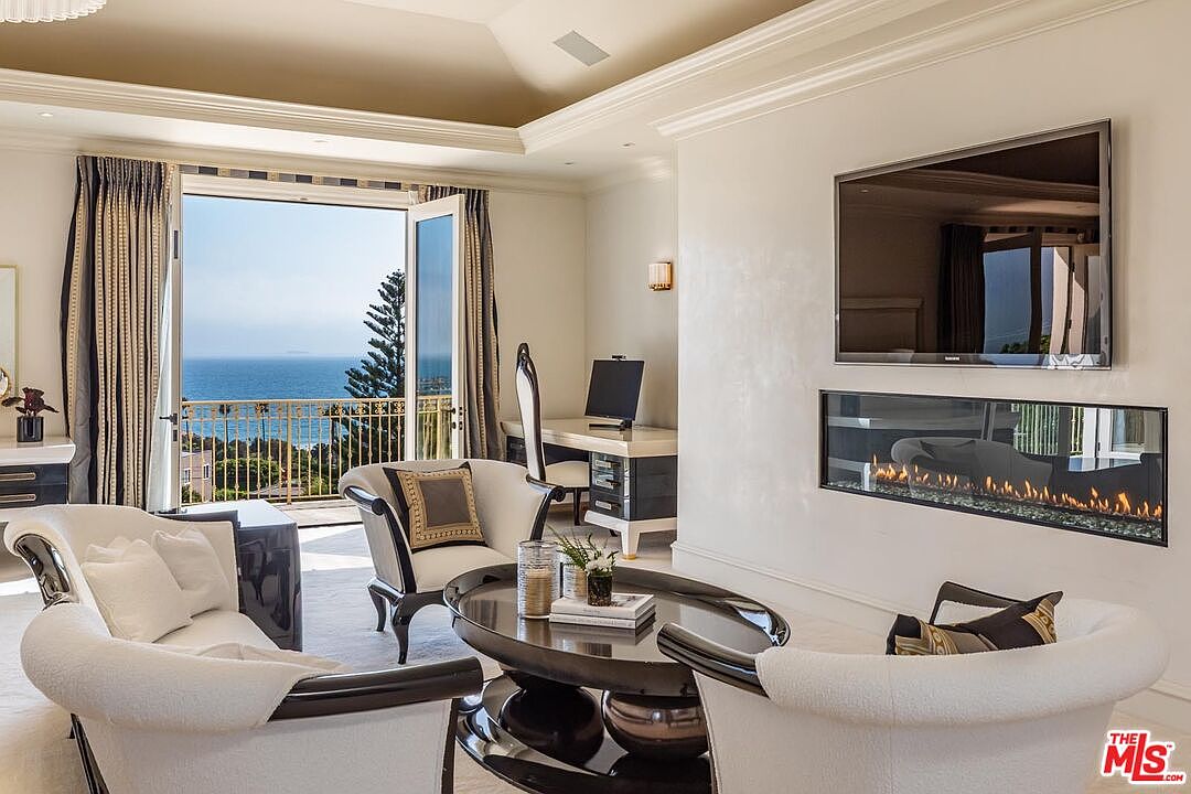 This sophisticated living room features a contemporary fireplace integrated into a sleek wall beneath a large flat-screen television. The space is furnished with elegant, curved white armchairs and a dark oval coffee table, all centered around a bright, airy atmosphere that opens onto a balcony with stunning ocean views. The room blends modern luxury with a serene coastal aesthetic, highlighted by high ceilings and refined architectural details.