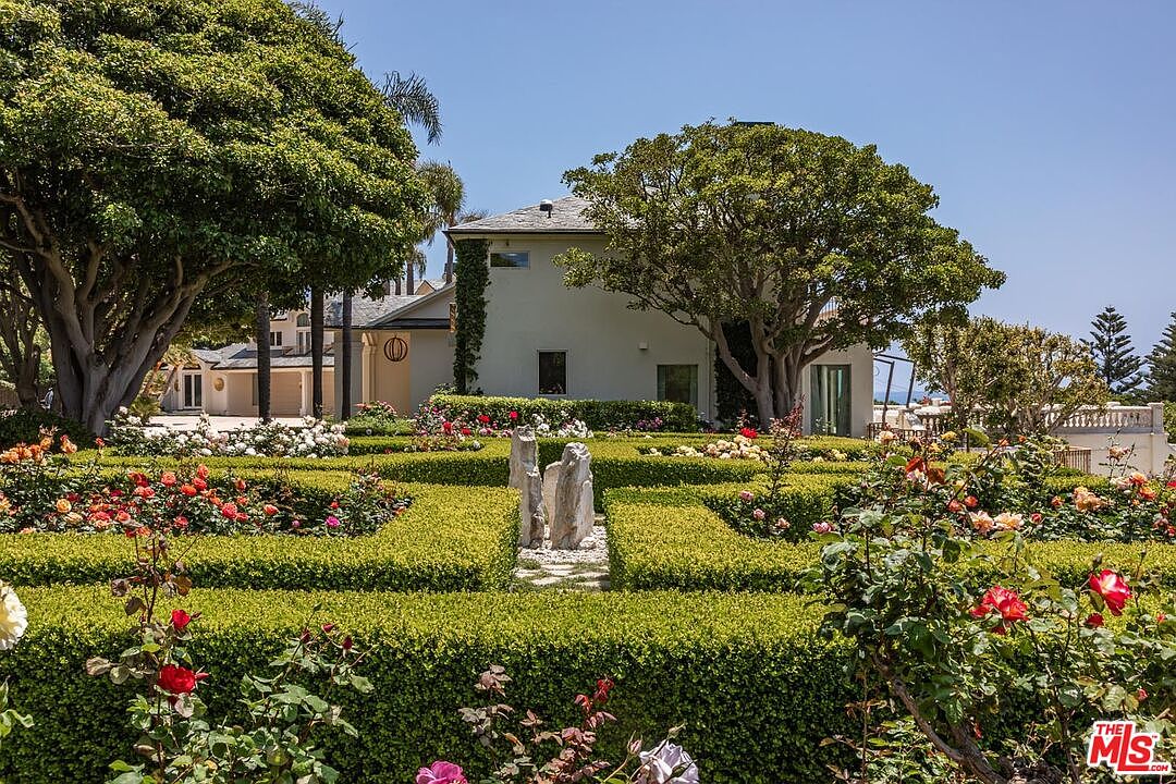 This image showcases a meticulously manicured formal garden featuring geometric boxwood hedges and vibrant rose bushes in full bloom. In the center, a striking stone sculpture serves as a focal point, leading the eye toward a stately, light-colored residence nestled behind mature trees. The perspective is a ground-level shot that emphasizes the lush, serene atmosphere of this expansive estate property.
