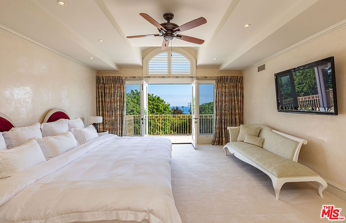 This spacious primary bedroom features a large, plush bed with white linens and a sophisticated cream-colored settee positioned against the wall. The room is anchored by a central ceiling fan and opens up to a private balcony through double doors, offering a stunning, unobstructed view of the ocean. The neutral color palette and elegant architectural details create a serene and luxurious atmosphere.