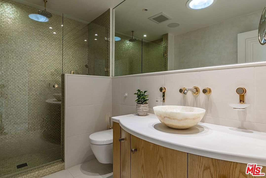 This modern bathroom features a sleek, curved wooden vanity topped with a unique, translucent stone vessel sink and gold-toned fixtures. A glass-enclosed walk-in shower with shimmering gold mosaic tiles serves as a luxurious focal point, complemented by a large wall-to-wall mirror that enhances the sense of space. The overall aesthetic is sophisticated and warm, blending natural textures with contemporary design elements.
