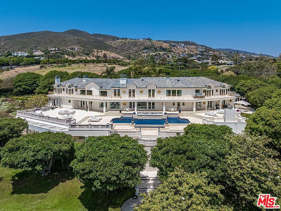 This stunning aerial view captures a sprawling, Mediterranean-style luxury estate set against a backdrop of rolling hills and a clear blue sky. The property features a massive multi-level terrace, a large infinity-edge swimming pool, and meticulously landscaped grounds surrounded by lush trees. The grand architecture and expansive outdoor living spaces convey an atmosphere of opulence, privacy, and high-end coastal living.