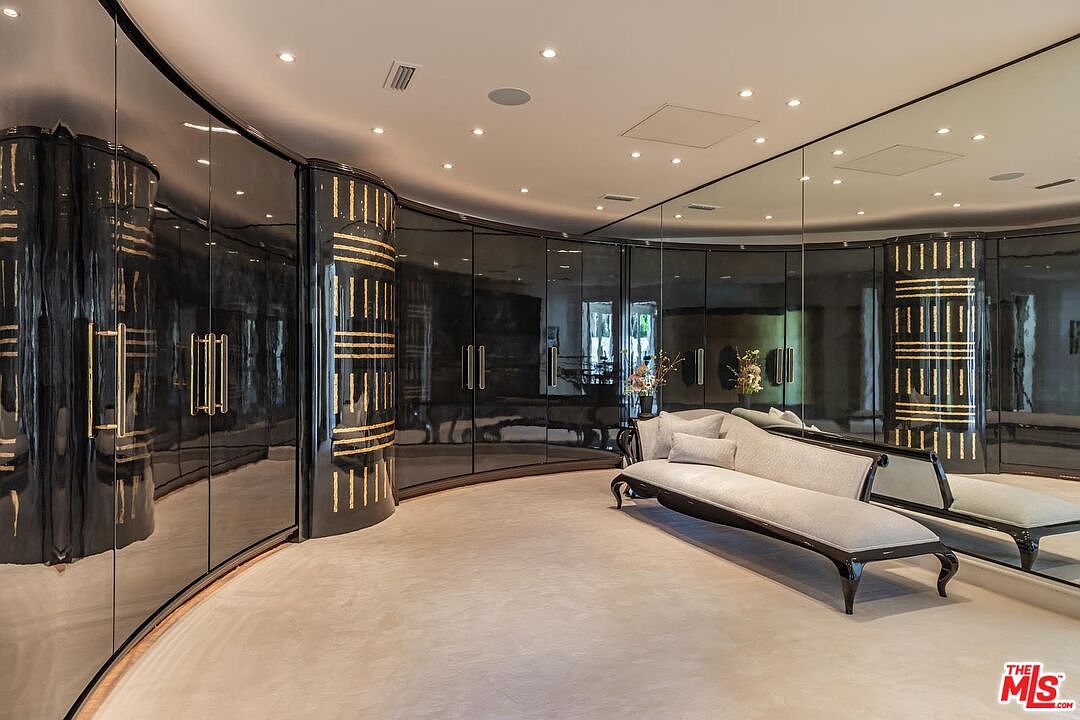 This luxurious, high-end walk-in closet features dramatic, curved black lacquer cabinetry with gold-accented detailing and integrated lighting. A sophisticated, light-toned chaise lounge sits in the center of the room, set against a backdrop of expansive floor-to-ceiling mirrors that enhance the sense of space. The overall aesthetic is one of opulence and refined, modern glamour.
