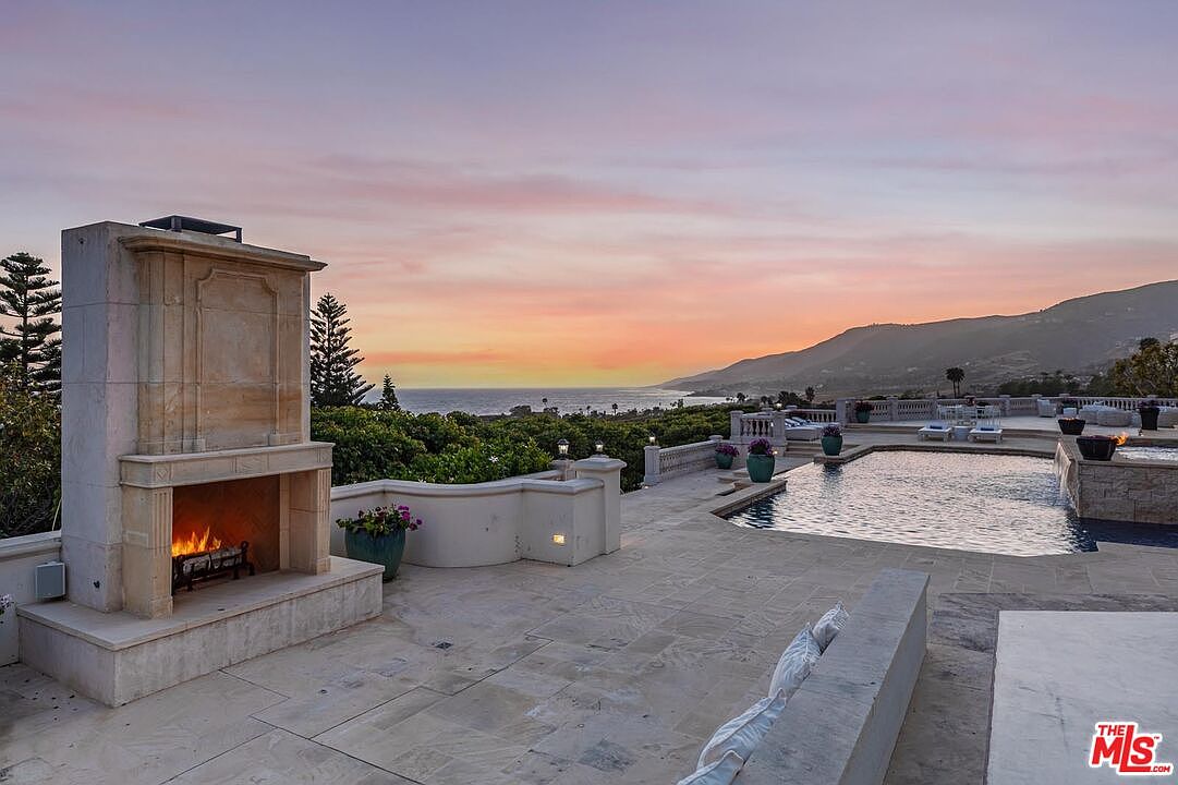 This luxurious outdoor terrace features a grand stone fireplace as a focal point, overlooking a stunning infinity-edge pool that blends seamlessly with the ocean horizon. The expansive stone patio is designed for high-end entertaining, offering panoramic sunset views over the coastline and mountains. The atmosphere is one of sophisticated relaxation, perfectly capturing the essence of coastal luxury living.