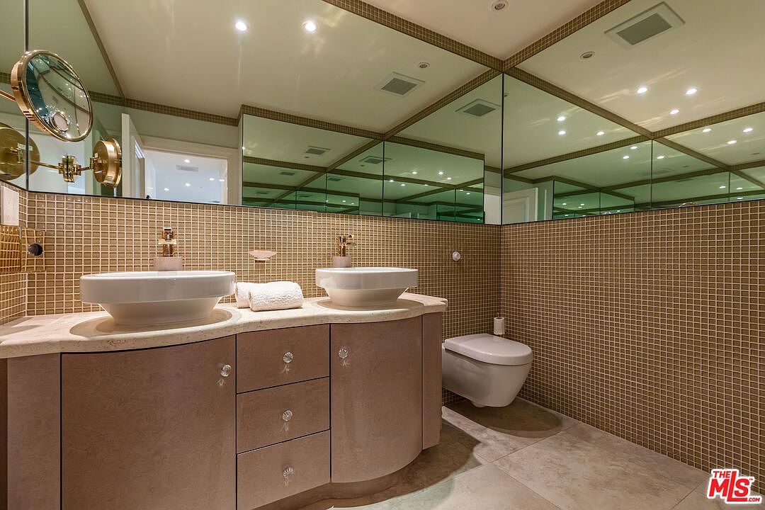 This primary bathroom features a double vanity with two white vessel sinks set atop a curved, light-toned cabinet base. The walls are adorned with small, square, metallic-toned mosaic tiles, complemented by extensive mirrored surfaces that create a sense of depth and brightness. A wall-mounted toilet is positioned to the side, and the overall aesthetic is contemporary with a touch of luxury.