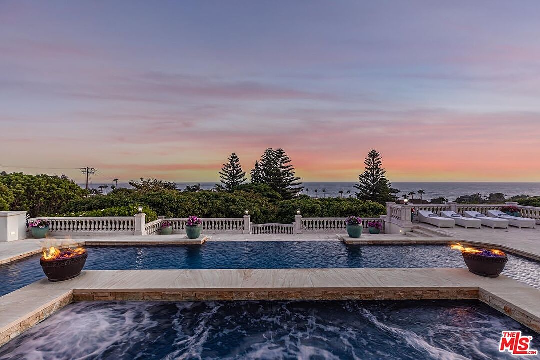 This stunning outdoor space features a luxurious infinity-edge pool and a bubbling spa, perfectly positioned to overlook a breathtaking ocean sunset. The stone-tiled deck is accented by elegant fire bowls and classic balustrade railings, creating a sophisticated and serene atmosphere. The panoramic view of the horizon and the lush greenery provides a high-end, resort-like feel ideal for relaxation and entertaining.
