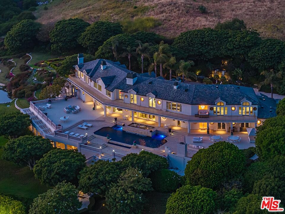 This stunning aerial perspective captures a sprawling, multi-level luxury estate nestled into a lush, hilly landscape at twilight. The residence features a grand architectural design with a slate roof, expansive stone terraces, and a beautifully illuminated swimming pool area that serves as the focal point of the backyard. Surrounded by mature trees and manicured grounds, the property exudes an atmosphere of exclusive privacy and sophisticated elegance.