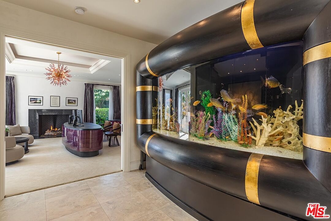This interior view features a prominent, custom-built, large-scale aquarium integrated into a dark, gold-accented wall, serving as a striking focal point in the hallway. Through the open doorway, a sophisticated home office is visible, complete with a dark wood desk, a fireplace, and a modern sputnik-style chandelier. The perspective captures the transition between the tiled hallway and the carpeted office, highlighting a luxurious and unique architectural design.