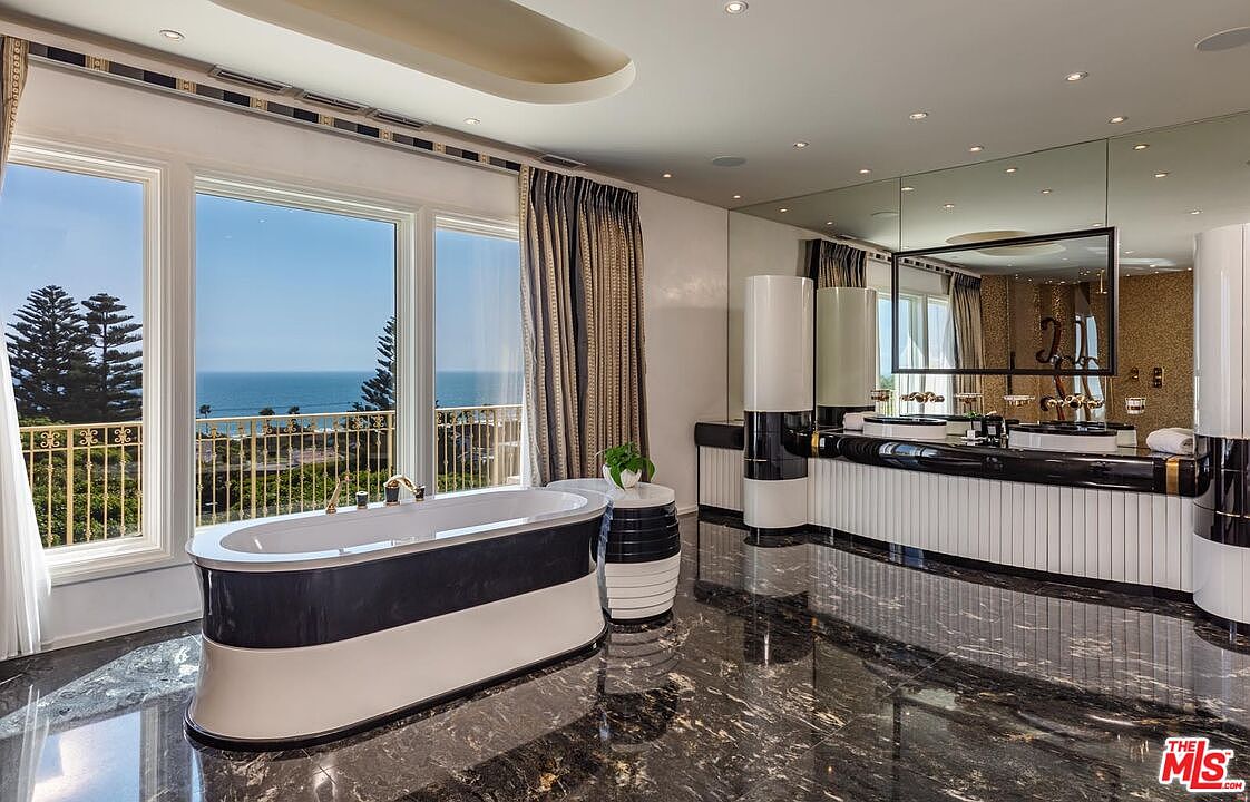 This luxurious primary bathroom features a striking black and white Art Deco-inspired aesthetic, highlighted by a sleek freestanding soaking tub and a matching double vanity. The room is anchored by polished black marble flooring with gold veining, while floor-to-ceiling windows offer a breathtaking, unobstructed view of the ocean. The space exudes opulence and sophistication, perfectly blending high-end materials with a serene coastal backdrop.