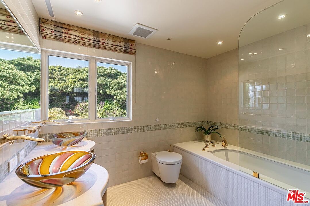 This bright and airy primary bathroom features a unique double vanity with colorful, artistic glass vessel sinks and gold-toned fixtures. The space includes a wall-mounted toilet and a deep soaking tub enclosed by a curved glass partition, all set against neutral, textured wall tiles. A large window provides ample natural light and a pleasant view of lush greenery outside, creating a serene and upscale atmosphere.