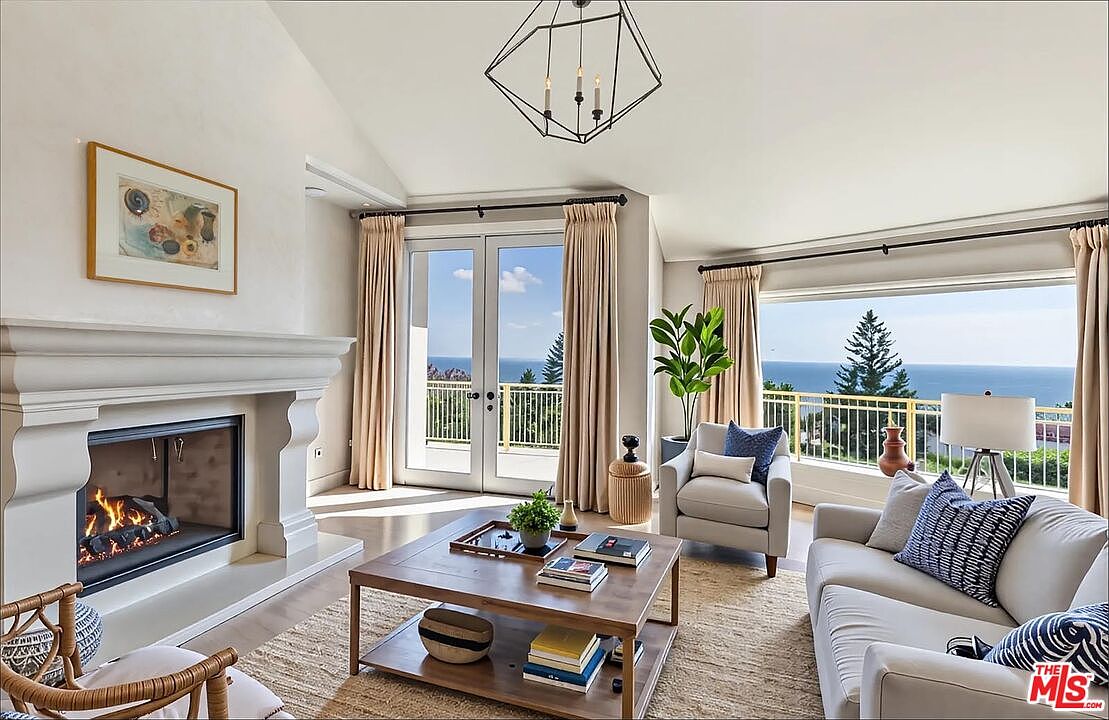 This bright and airy living room features a sophisticated coastal aesthetic, highlighted by a grand fireplace and expansive windows that frame a stunning ocean view. The space is furnished with a comfortable sofa, a plush armchair, and a central wooden coffee table, all resting on a textured jute rug. The vaulted ceiling and geometric chandelier add a touch of modern elegance, creating a serene and inviting atmosphere perfect for relaxation.