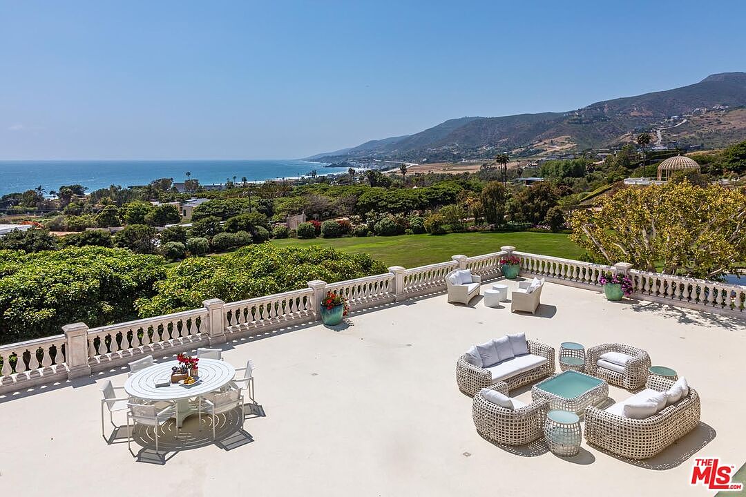 This expansive, elevated stone terrace offers a breathtaking panoramic view of the coastline, lush green hills, and the Pacific Ocean. The space is elegantly furnished with a white dining set and a comfortable wicker lounge area, all framed by a classic balustrade railing. The scene captures a serene, luxurious outdoor living experience perfect for entertaining while overlooking the vast landscape.