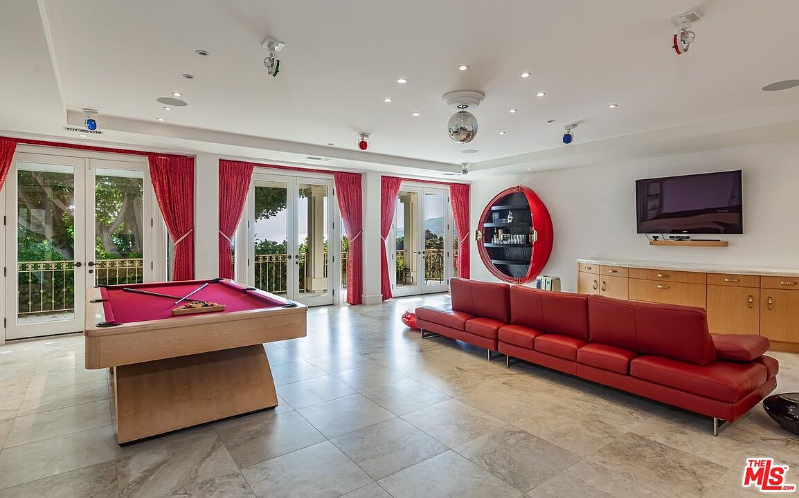 This spacious game room features a vibrant red leather sectional sofa and a matching pool table, creating a bold and energetic entertainment space. The room is accented by a unique circular wall-mounted bar unit, recessed ceiling lighting, and multiple sets of French doors that open to an outdoor balcony. The polished tile flooring and bright white walls provide a clean, modern backdrop for this lively recreational area.