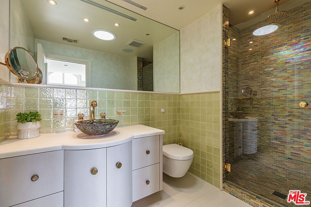 This vibrant bathroom features a unique, multi-colored glass vessel sink set atop a curved white vanity with gold hardware. The walls are adorned with light green square tiles, while the walk-in shower showcases a striking, iridescent mosaic tile pattern. A large, frameless mirror reflects the space, enhancing the bright and eclectic aesthetic of this well-appointed room.