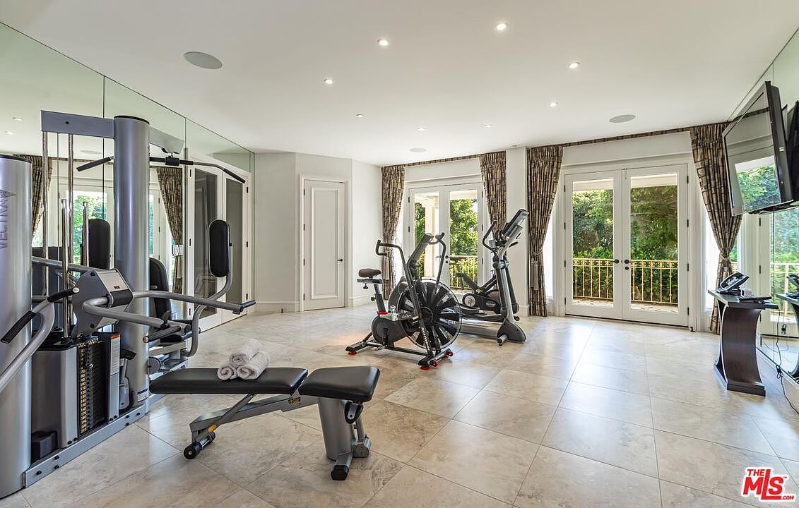 This spacious home gym features light-colored tile flooring, mirrored walls, and multiple sets of French doors that open to a balcony, allowing for ample natural light. The room is equipped with a professional-grade multi-station weight machine, an air bike, and an elliptical trainer, creating a comprehensive fitness environment. The overall aesthetic is clean, bright, and functional, designed for a high-end residential property.