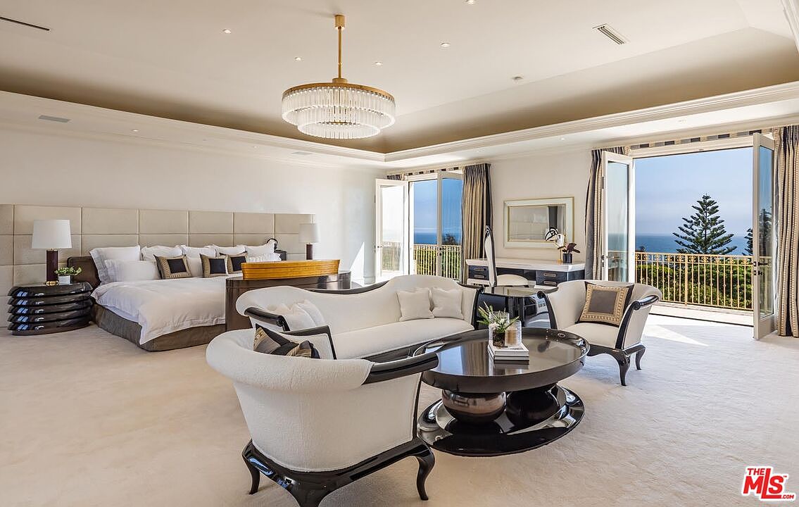 This luxurious primary bedroom features a spacious layout with a plush king-sized bed, a sophisticated seating area with a curved sofa, and a central coffee table. The room is elegantly appointed with a crystal chandelier, neutral tones, and expansive glass doors that open to a balcony with stunning ocean views. The overall aesthetic is one of refined opulence and serene comfort, perfectly suited for a high-end estate.