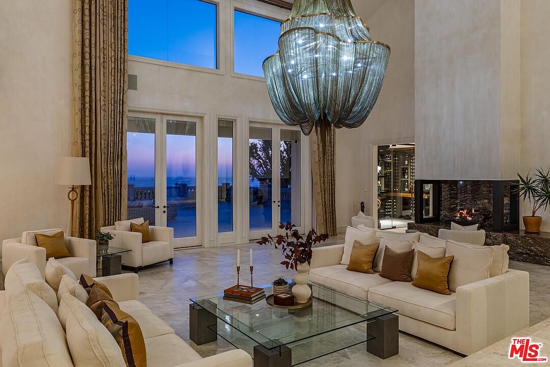 This expansive, high-ceilinged living room exudes luxury with its neutral-toned plush sofas, a large glass-topped coffee table, and a striking, oversized metallic chandelier as the focal point. A stone-clad fireplace adds warmth to the space, while floor-to-ceiling windows offer a breathtaking view of the ocean at dusk. The room is elegantly furnished with a mix of contemporary and classic elements, creating a sophisticated and inviting atmosphere.